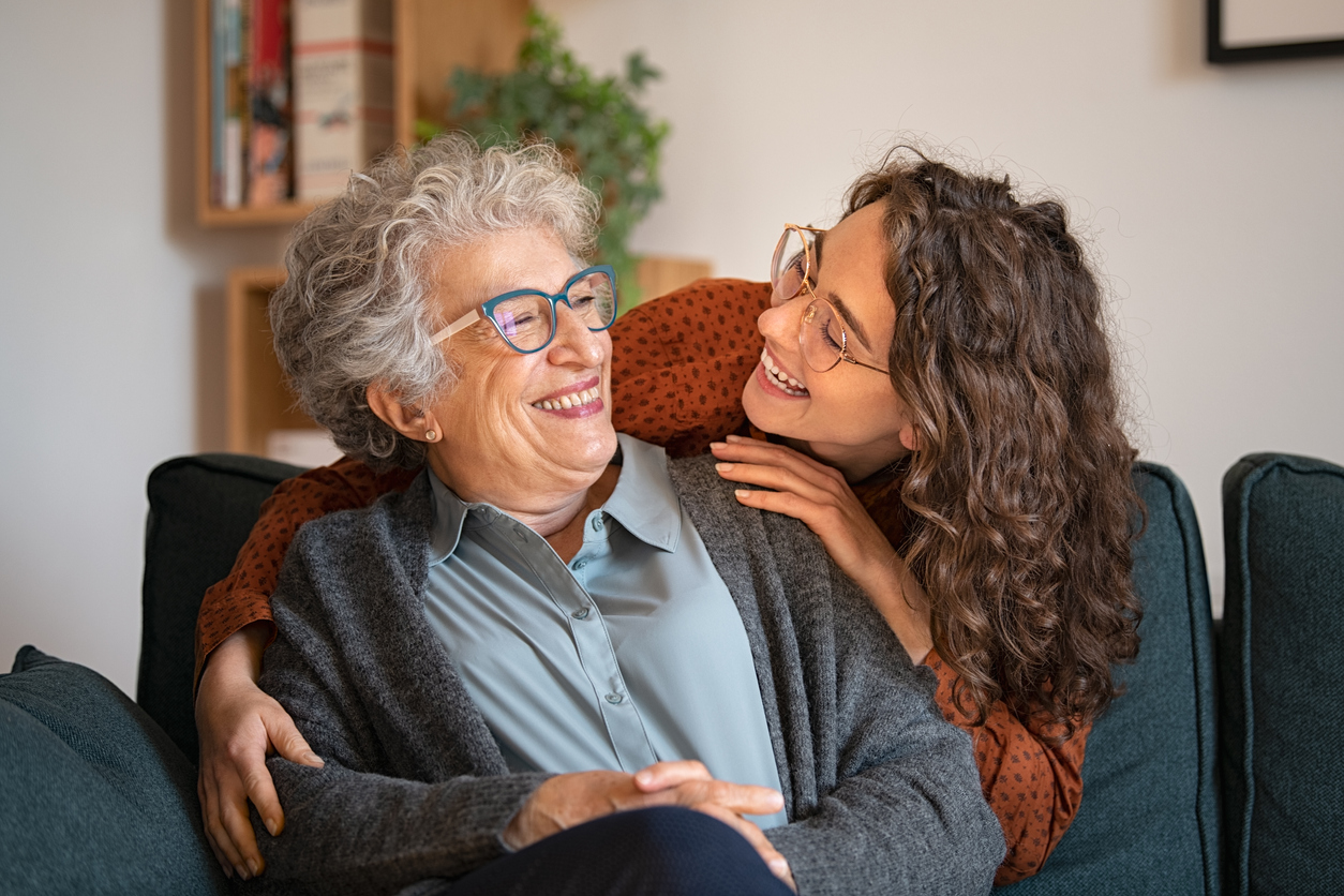 Elderly woman and younger adult woman smiling and sitting close together on a couch.