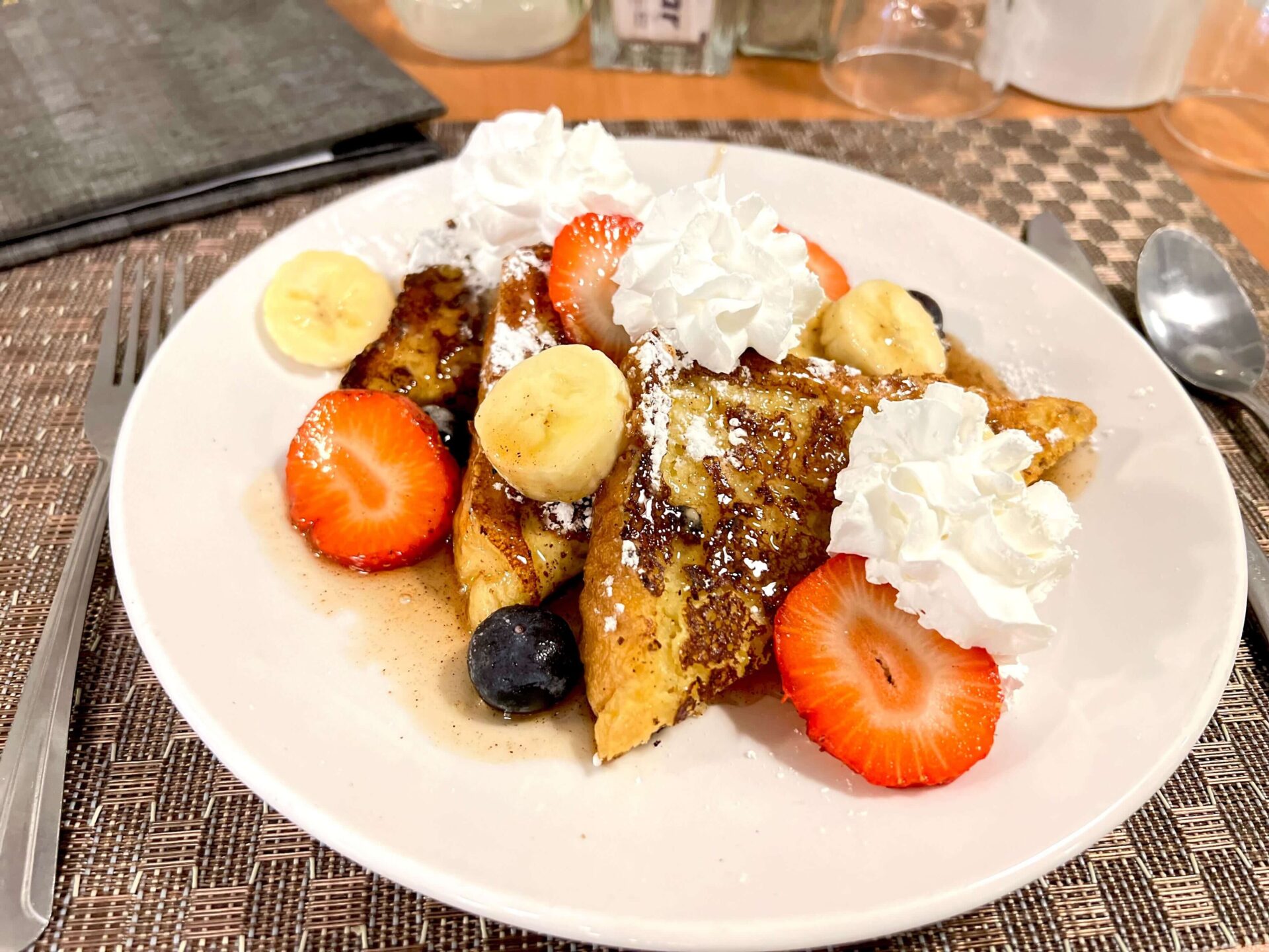 French toast with whipped cream, strawberries, blueberries, and banana slices on a white plate.