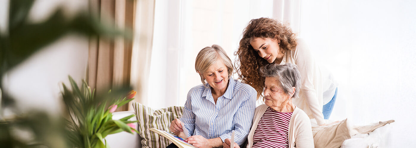 Two elderly women sit on couch and review a photo album while younger woman looks on