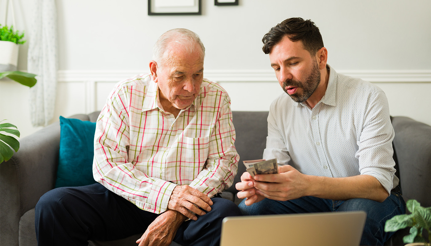 Senior man and younger man sit on a couch together and review finances